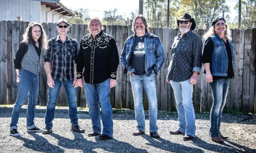 Group of older white men wearing jeans and flannel or button-down shirts standing outside in front of a fence 