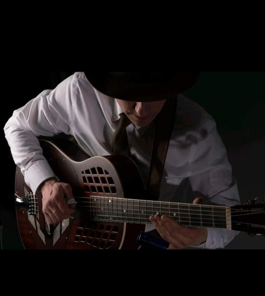 A man with a wide brimmed black hat covering his face. He is looking down toward a guitar he's holding.