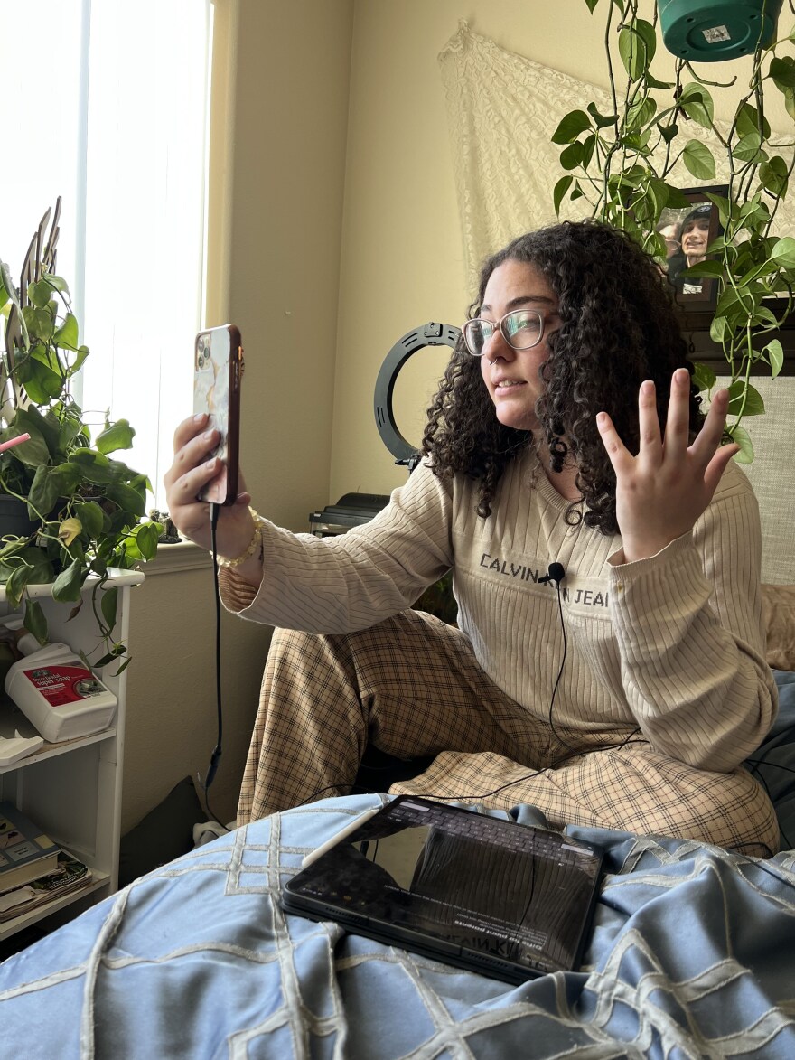 A woman is sitting on her bed and holding her phone up to her face while talking with her other hand. She’s brightly lit because she’s sitting in front of a window. There’s a ring light and a plant with several feet-long vines behind her.