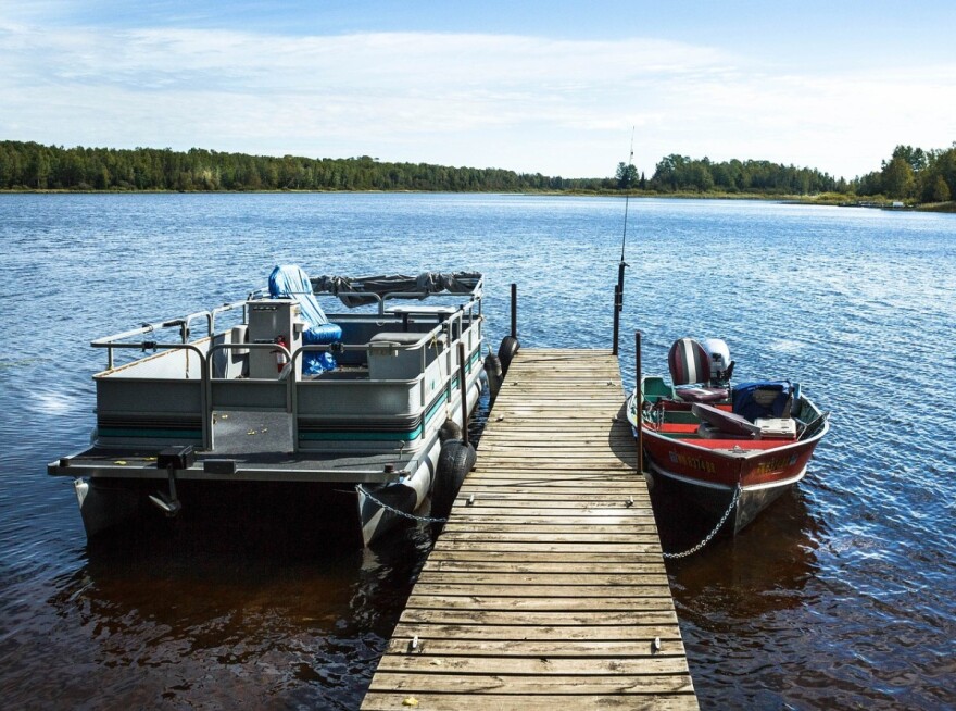  A pontoon boat and a red fishing boat are parked at a dock in a lake. A slight breeze ripples the water and green trees are visible in the background