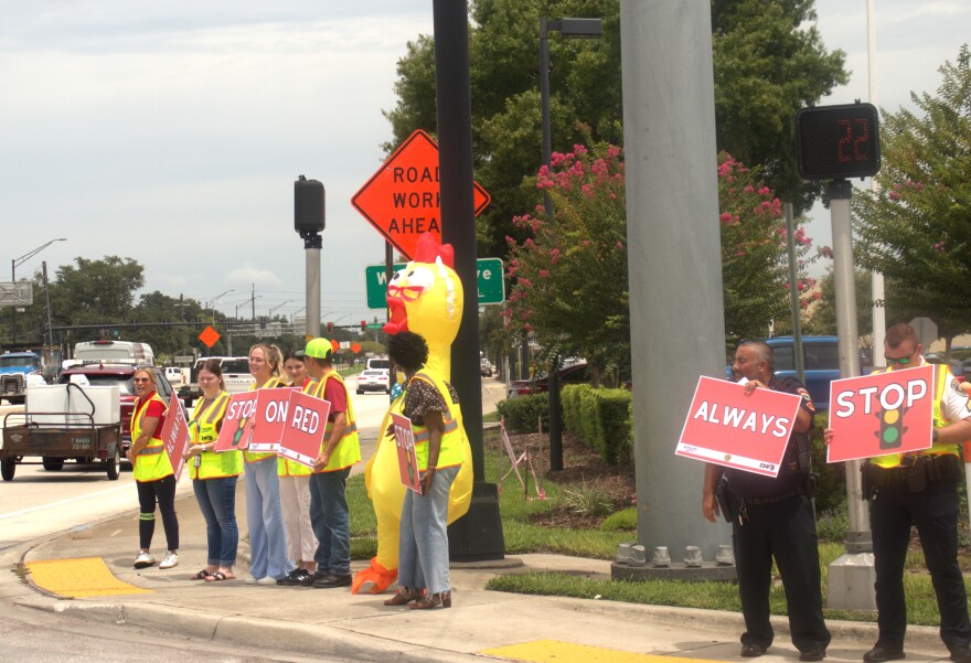 A group of individuals in neon traffic vests — plus, one in an inflatable chicken suit —  hold red signs that spell out "Always Stop On Red." They're standing at an intersection; an orange "road work ahead" sign and timed crosswalk light are visible, along with a line of cars. 