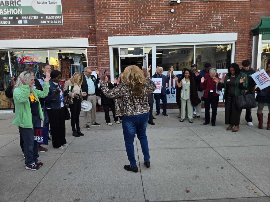 A group of people stand in a semi-circle on a concrete sidewalk in front of a brick building storefront, raising their hands toward a woman with long, curly blonde hair who is facing away from the camera. The diverse crowd of onlookers includes individuals holding signs that read "Mike Rogers for U.S. Senate." 