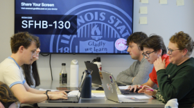 Students use laptops while they sit together at a tall table.