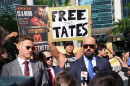 Two suited men speak on microphones while men behind them hold up signs. 
