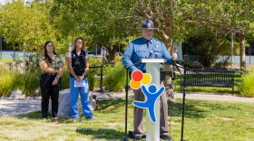 A man in a state trooper uniform stands at a podium, addressing an audience. Two women stand behind him, looking on.
