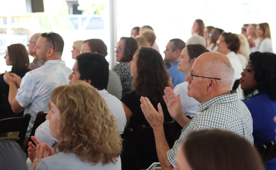 Audience members sitting closely in a brightly lit space under a tent, attentively clapping. 