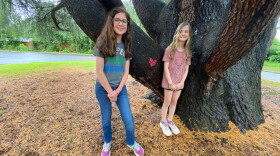 Park Road Montessori students Celia Kaul (left) and Ada Lee Smith stand at the base of the deodar cedar they're trying to save.