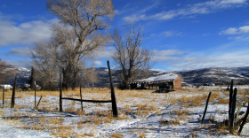 An old, worn-down shed sits in a field with yellow grass and snow. There are old wood fences in the foreground and a mountain range in the background.