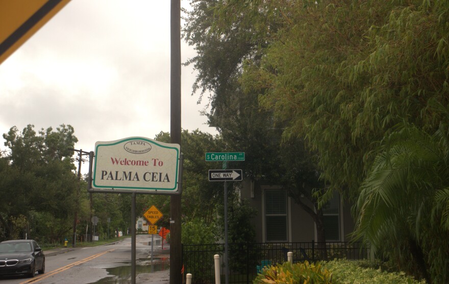 A vintage sign reads "Welcome to PALMA CEIA" on a street corner. Lush greenery, one house and one car are visible in the background.