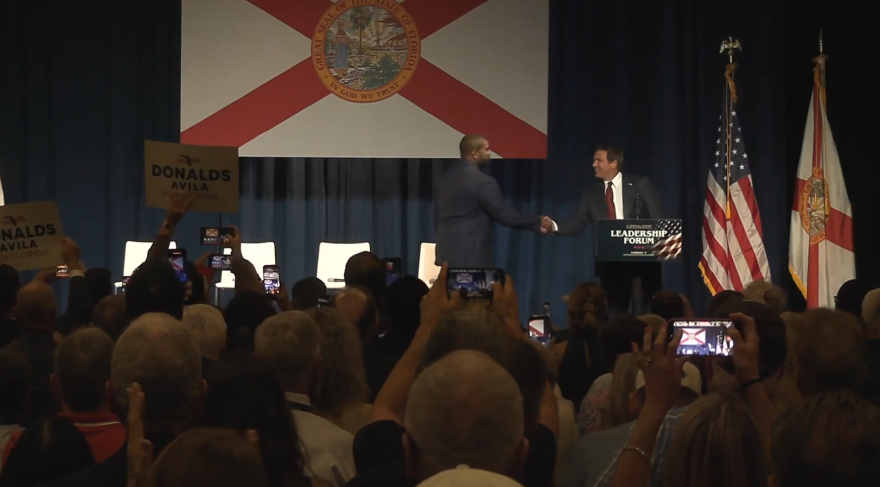 Florida Gov. Ron DeSantis shakes hands with U.S. Rep. Byron Donalds onstage in front of a large Florida flag as audience members watch and take photos.