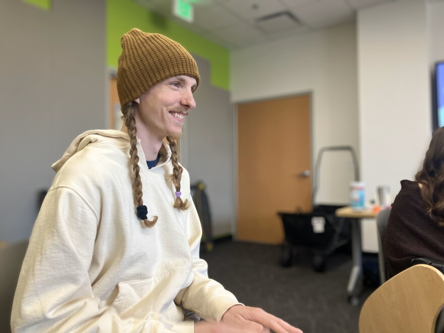 A man with long hair and two braids, wearing a brown knitted hat, smiles as he has his hands on a drum.