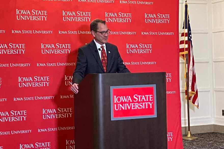 A man in a dark suit stands at a podium in front of a backdrop that says Iowa State University.