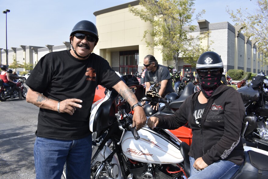 A man throws up a peace sign and poses next to his bike. There is a woman next to him wearing a helmet and mask.