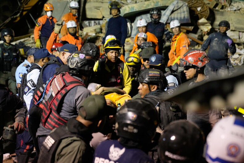 A group of people wearing helmets and other equipment carry a person through the rubble of a building. 