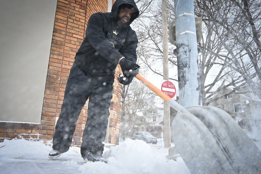 A man wearing alpine coat and pants uses a snow shovel to clear a sidewalk. Behind him is the corner of a brick building and on the other side of the street are row houses.