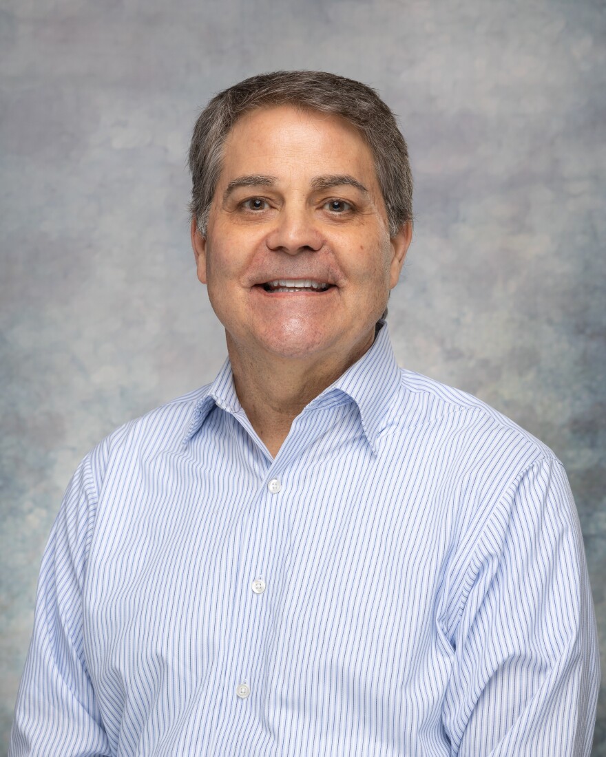 Portrait of Mike Downhour, president and CEO of the Monroe Chamber of Commerce, wearing a light blue striped button-down shirt against a neutral studio background.