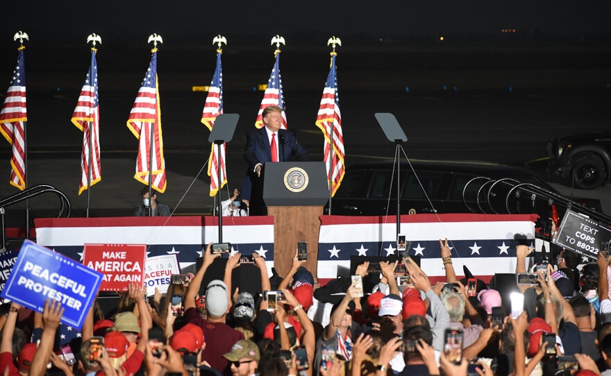 Afuera está oscuro. Un hombre se para en un podio con cinco banderas estadounidenses detrás de él. Está frente a una multitud.