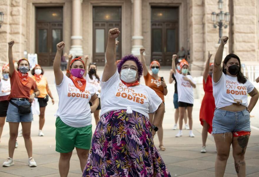 A group of women stand in formation in front of the Texas Capitol with a fist raised in the air. Some are wearing shirts saying "Bans Off Our Bodies".