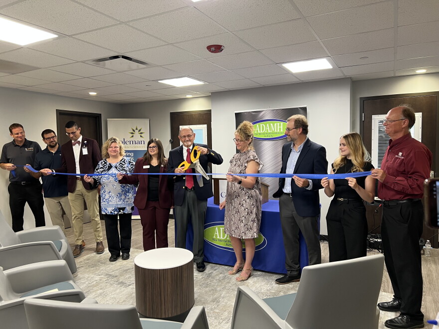 A man holding a large pair of ceremonial scissors cuts a ribbon held by multiple people to open a new building. 