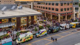 Exterior view of Lenexa Public Market, with food trucks lining the road in front of the brick and glass building, and people on the sidewalk around the building. 