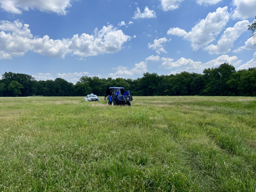  A white pickup truck follows a blue tractor through a pasture of green grass. Behind them, a line of trees and a blue sky with fluffy white clouds.