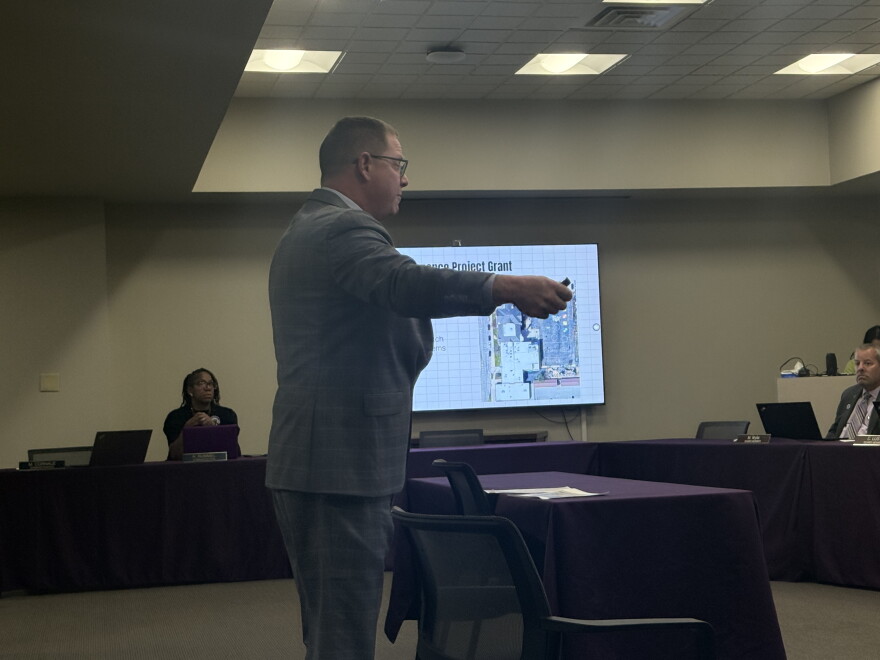 A man in a gray suit stands and points a remote toward a screen showing a slide titled “School Maintenance Project Grant,” with an aerial photo of a school building, as board members watch from a purple-draped table.