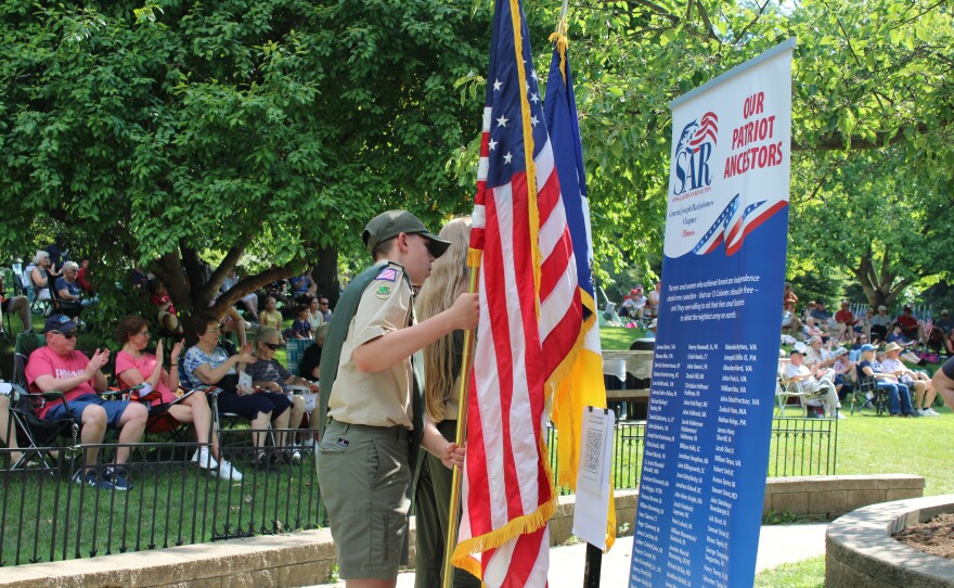 A Boy Scout in uniform holds an American flag beside a Sons of the American Revolution banner listing the names of local Revolutionary War patriot ancestors, with a seated audience behind him.