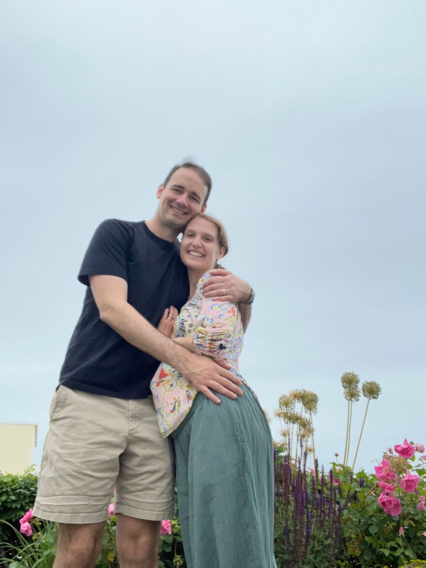 Lindsey Huebner and her husband Eric during Lindsey's pregnancy with Noah. Lindsey is a blonde woman in a green dress and multicolored shirt. Her husband Eric is wearing a dark t shirt and khaki shorts. They are standing in front of a bed of flowers. 