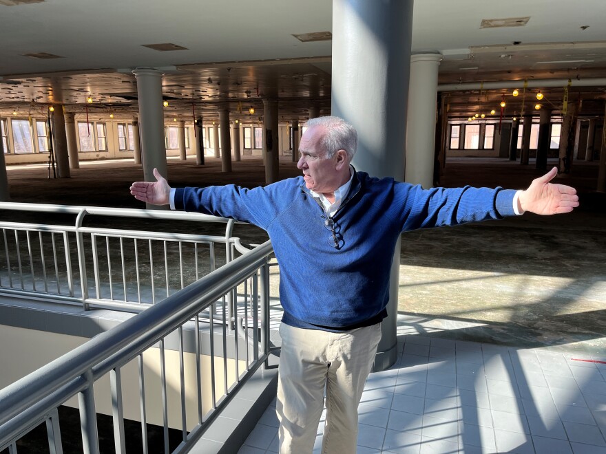  Ken Greene, director of commercial development for Sibley Square, gestures with arms wide, explaining how the fifth-floor of Sibley Square will be divided up for the future offices of Bandwidth, other common areas and still-unleased space. The sun is shining in through a large, glass atrium room. He is standing alongside the railing. 