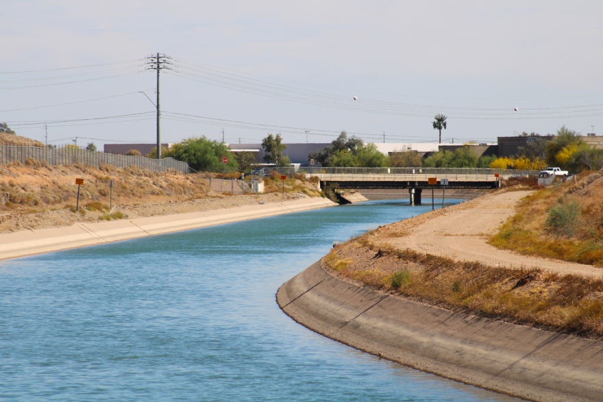 Water flows through a canal in an arid area. There's a dirt path running along it on one side and a small bridge up ahead.
