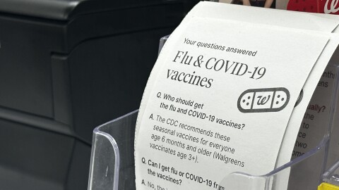 A stack of handout sit in a clear container next to a check-out station at a Walgreens pharmacy counter. The top of the handout reads "Your questions answered" in small font, followed by "Flu and COVID-19 vaccines." The handout answers several questions, including "Who should get the flu and COVID-19 vaccines?" and "Can I get flu or COVID-19 from the vaccines?" 