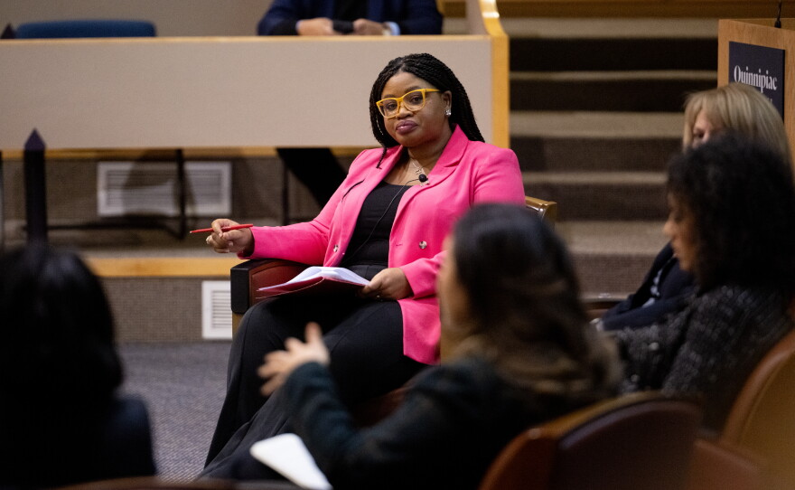 Khalilah Brown-Dean wears a pink jacket and is the only one facing the camera as she speaks with a group of women panelists seated at the front of a classroom.