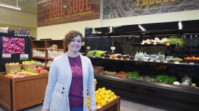 A woman in a light gray knitted coat, purple shirt and blue jeans stands in the produce section of a grocery store. She faces towards the camera with a small smile, leaning against a display of yellow lemons and other produce. Behind her can be seen one other display filled with apples, along with a larger row of vegetables in a black shelf against the wall. 