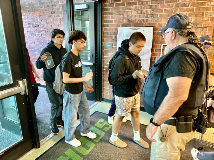 Three students enter through the doors of a school and show their phones and IDs to a security officer.