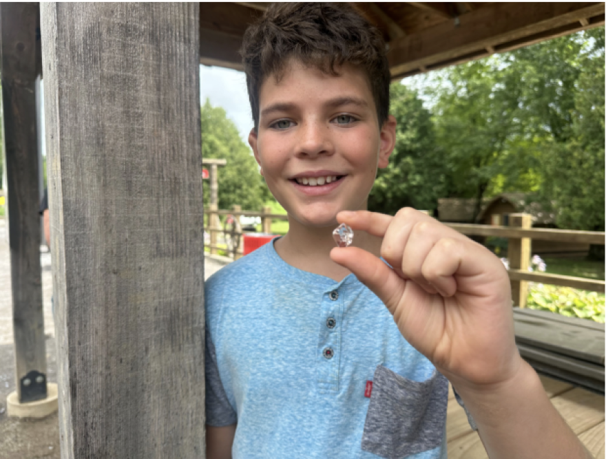 A boy smiling in a blue shirt holds a clear faceted crystal between his fingers.