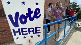 A large "Vote Here" sign is posted along the rails of a concrete ramp leading to a door where people are lined up to vote.