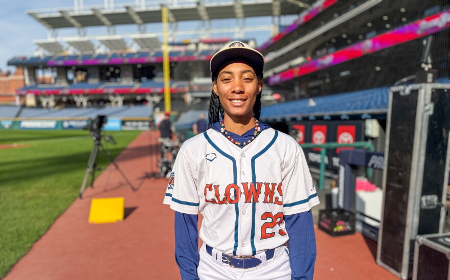 A Black woman baseball player dressed in an Indianapolis Clowns uniform smiles for the camera on the field of Progressive Field.