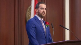 Man  with a groomed full beard in a blue suit stands behind a podium