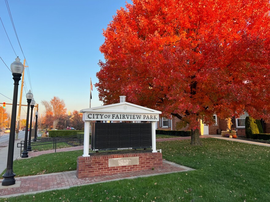 A sign reading City of Fairview Park on a green lawn next to a blazing red tree in autumn.