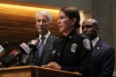 A woman in a police uniform stands at a podium. Two men stand behind her. 
