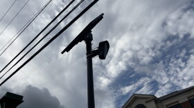 A Flock camera against a cloudy sky, with power lines and a brick building in the background.
