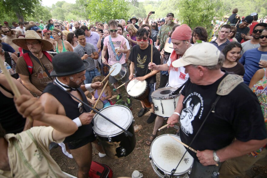 Several people play portable drums outdoors while a crowd watches.