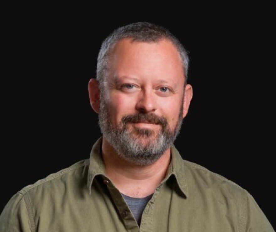 A man with a graying beard looking at a camera with a black background behind him.