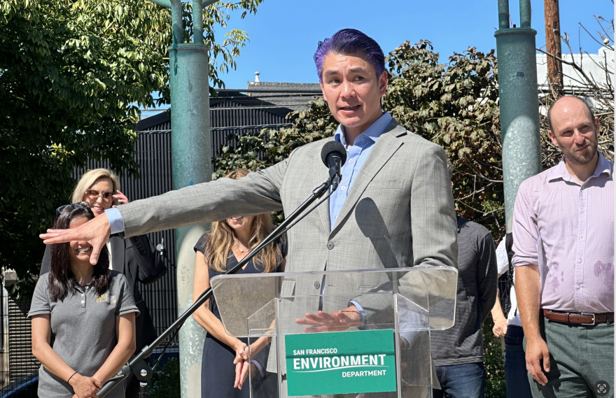 A man in a suit with purple hair stands in front of a transparent podium with an San Francisco Environment Department placard. He gestures to the group of people standing behind him.