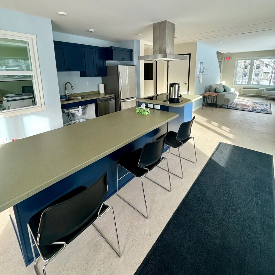 Interior space with a long table, three black chairs, kitchen and living room areas in the background