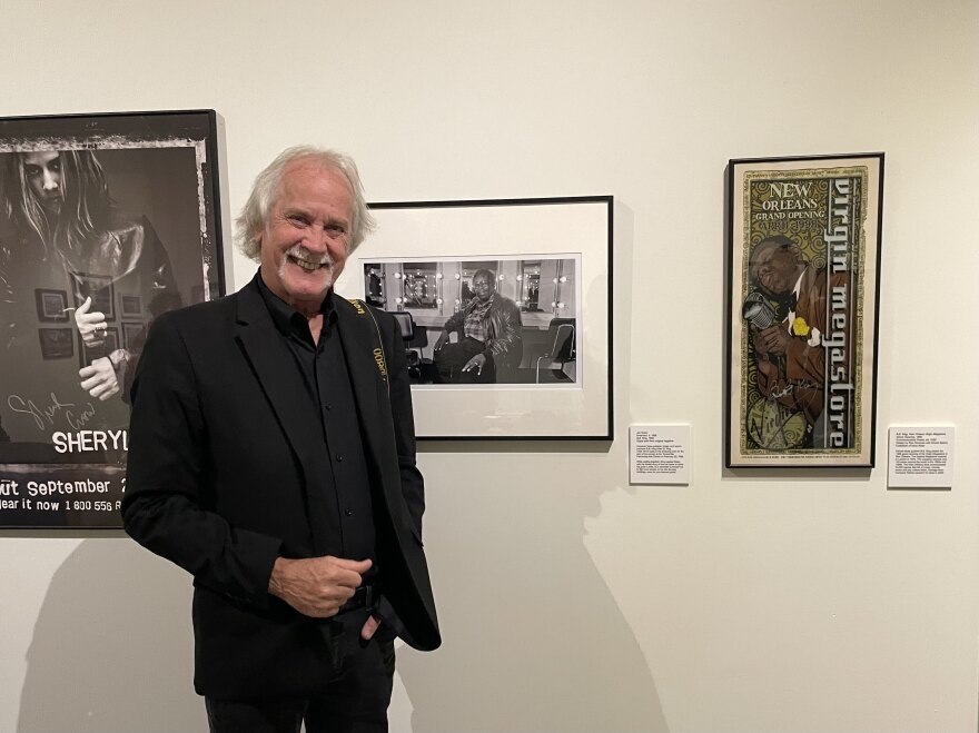 A grey haired man in a black suit holding a camera stands in front of a black and white photo of blues musician BB King. There are posters to the right and left of him.