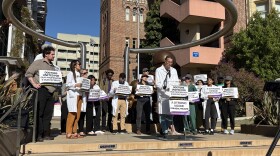An individual stands before a mic and podium in a doctor's white coat speaking, with a small crowd of other medical staff members standing behind the speaker holding up signs in favor of unionizing. 