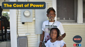 An older Black man with a gray goatee standing on the side of a yellow house, with his left hand on a young girl's shoulder. They're standing in front of a power meter on the house. A grey and yellow text saying "The Cost of Power" to the left 