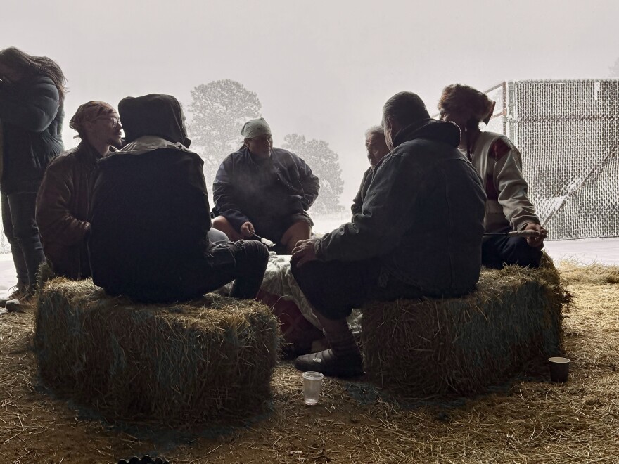 Six Native American drummers sit on hay stacks in a circle with a large drum in the middle and a snow scene in the background.