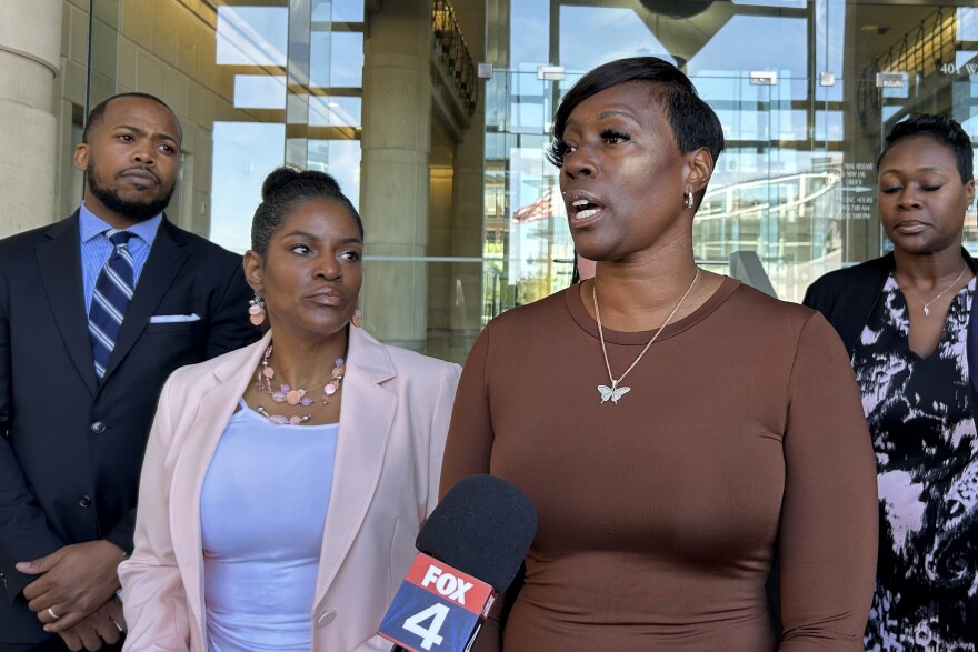 A photo of Crystal Mason, a Black woman with short, straight black hair, wearing a brown dress and a butterfly necklace. She speaks into a microphone at a press conference, surrounded by her attorneys and supporters.